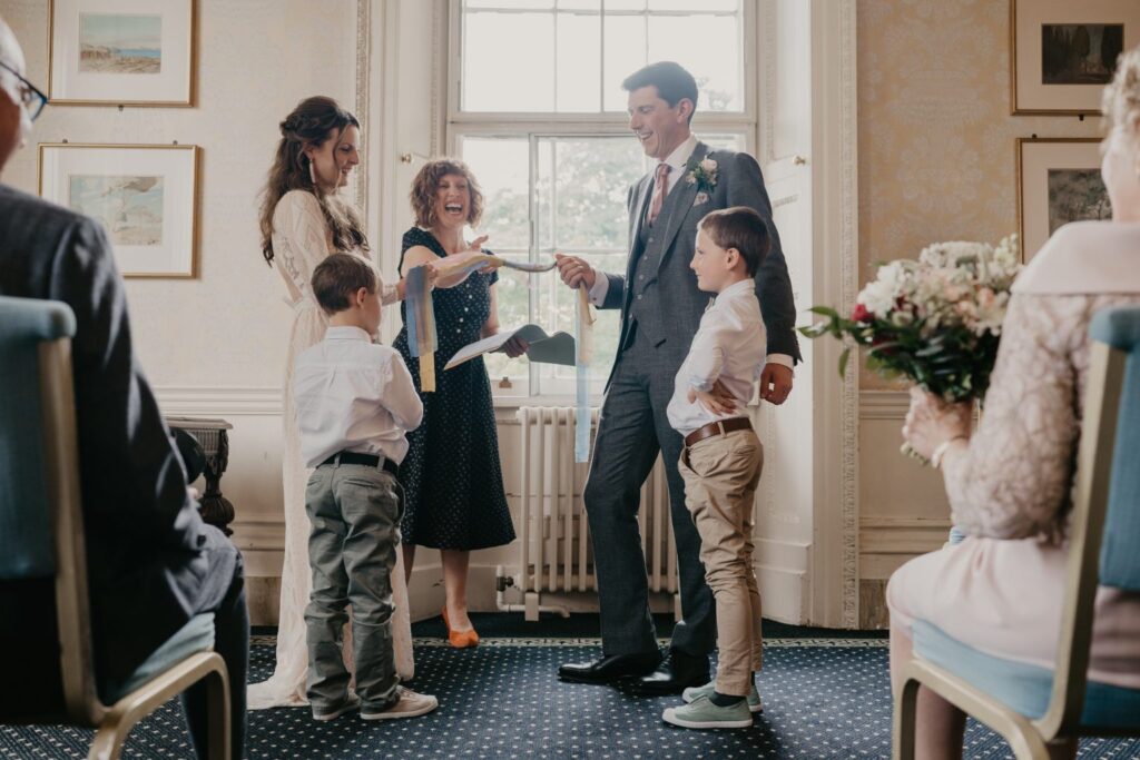 Kim Williams Wedding Indoor wedding ceremony with celebrant, Amy Lee, and family, including children participating, in a well-lit room with large windows.