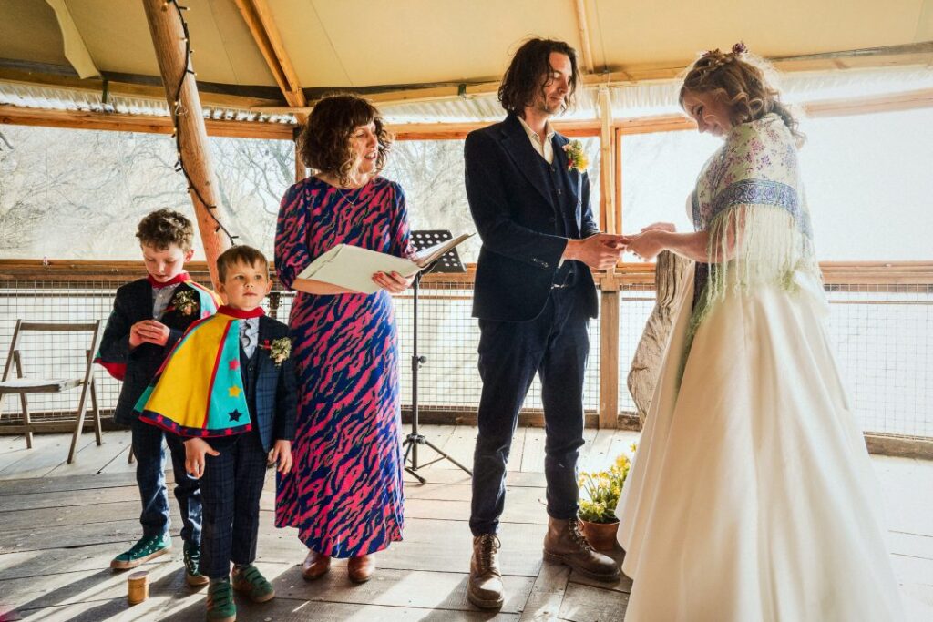 Matt Parry Photography A celebrant in a colorful patterned dress stands beside a couple exchanging vows. The groom, wearing a dark suit, holds hands with the bride, who is dressed in a white gown. A young child, wearing a vibrant outfit with a colorful cape, stands near the celebrant, watching the ceremony. The setting has a rustic feel, with wooden beams and natural light coming through large windows.