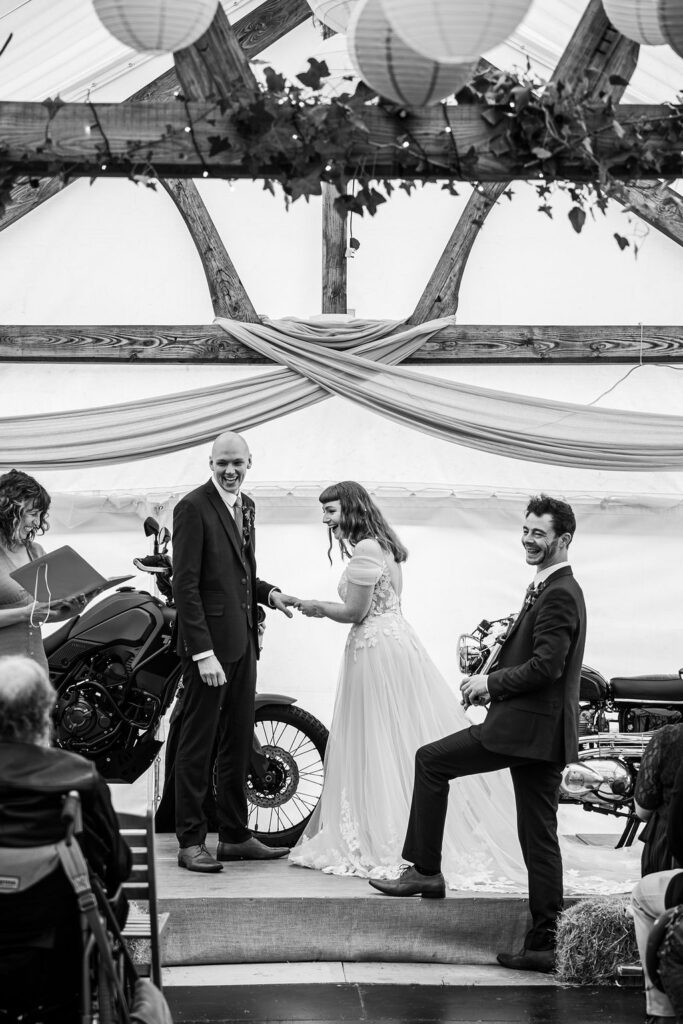 A D photography Black and white image of bride and groom holding hands, with motorcycles in the background under a wooden structure. Amy lee, the celebrant, smiles next to them.