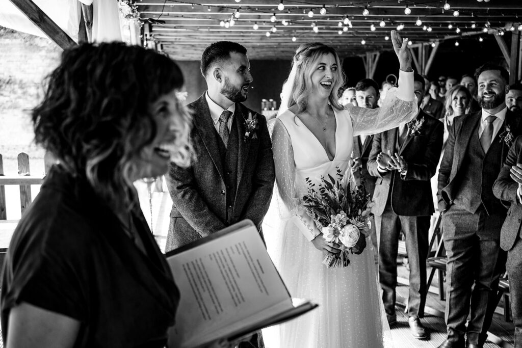 Lovell Photography Black and white image of bride and groom standing together during their ceremony, with celebrant, Amy Lee, holding an open book