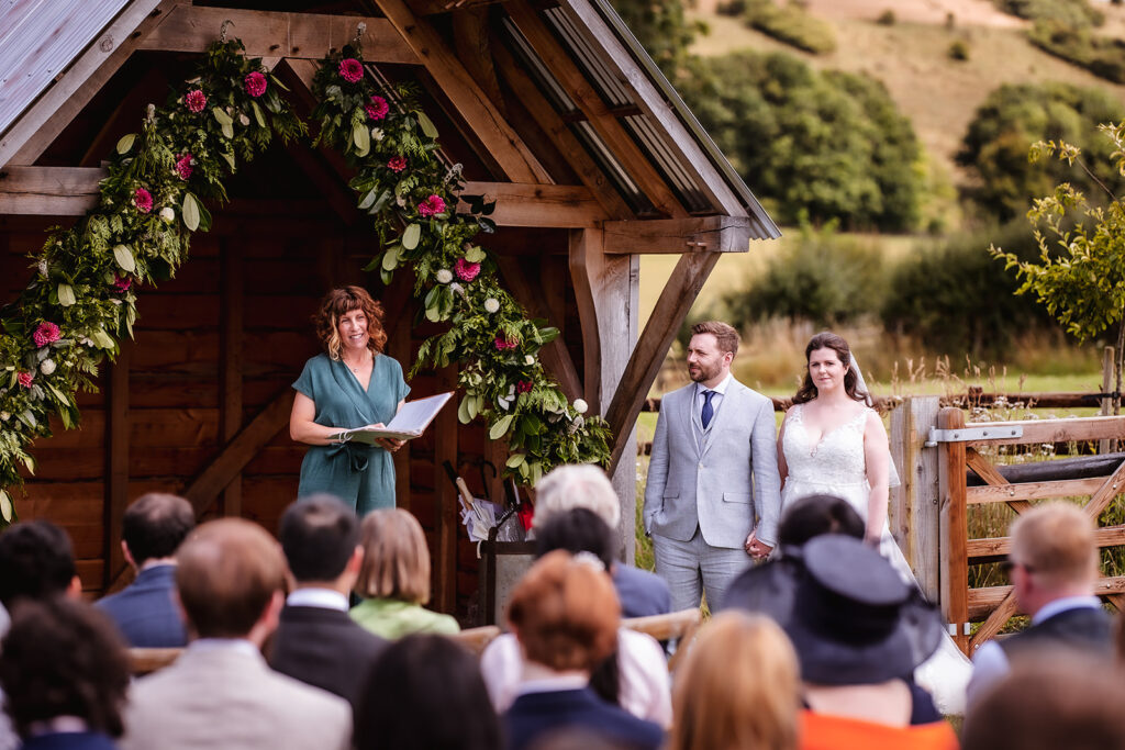 Adela Photography Bride and groom holding hands in front of a small wooden chapel adorned with flowers, with celebrant, Amy Lee, smiling at the couple and guests looking on.
